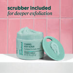 Open jar of Onyx Professional Foaming Body Scrub with scrubber lid leaning upright in front of pink tile. Text reads ‘scrubber included for deeper exfoliation.